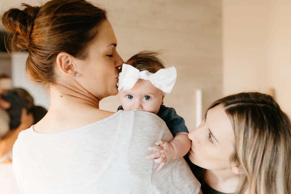 Who We Are 8 Two young lesbian mothers holding and kissing their infant daughter. Baby is wearing a large bow and making eye contact with camera.
